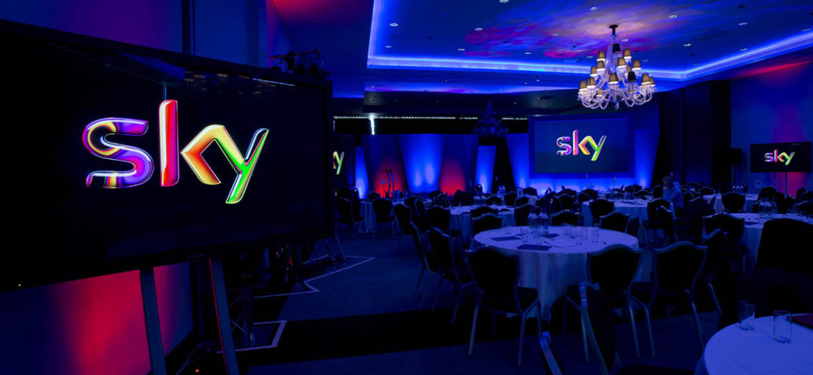Sky Technology Division Conference showing screens with the Sky logo in a conference venue with round tables and coloured lighting effects.