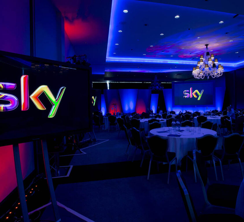 Sky Technology Division Conference showing screens with the Sky logo in a conference venue with round tables and coloured lighting effects.