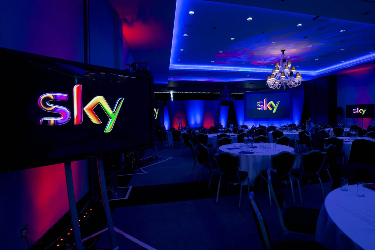 Sky Technology Division Conference showing screens with the Sky logo in a conference venue with round tables and coloured lighting effects.