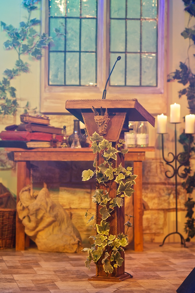 An example of conference stage design featuring a lectern with ivy wrapped around the base, and a table of books, candelbra and mock window behind.