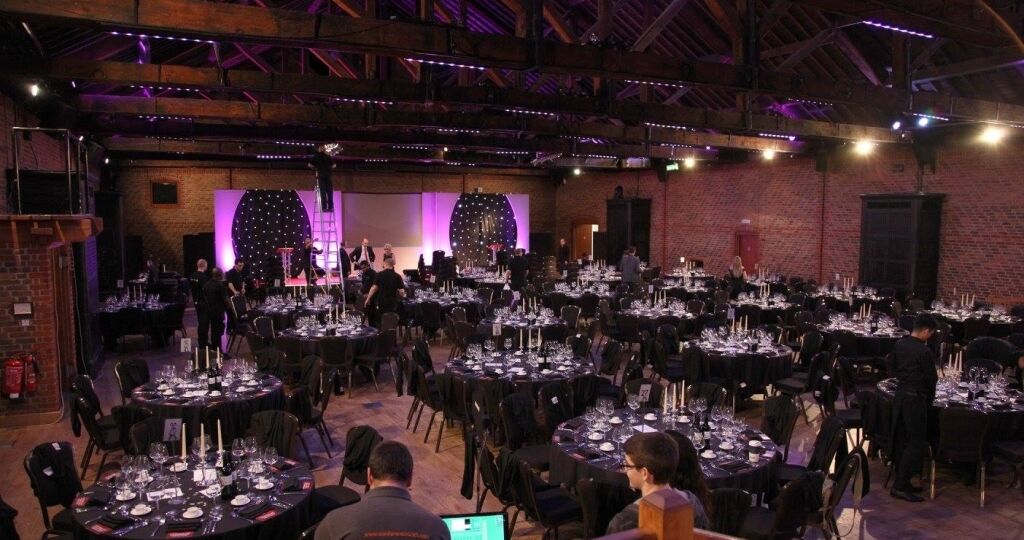 A large hall set up with round tables with chairs and candles, a stage and staff involved with corporate event production at 'The Wow Awards'.