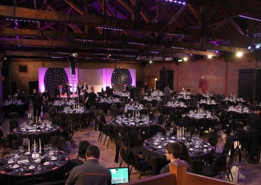 A large hall set up with round tables with chairs and candles, a stage and staff involved with corporate event production at 'The Wow Awards'.