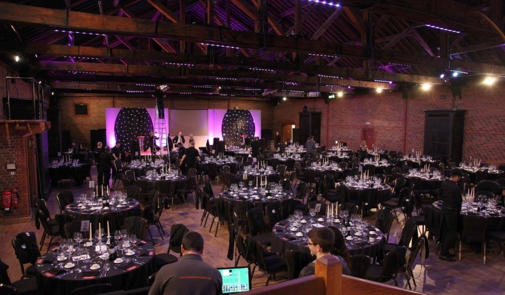 A large hall set up with round tables with chairs and candles, a stage and staff involved with corporate event production at 'The Wow Awards'.