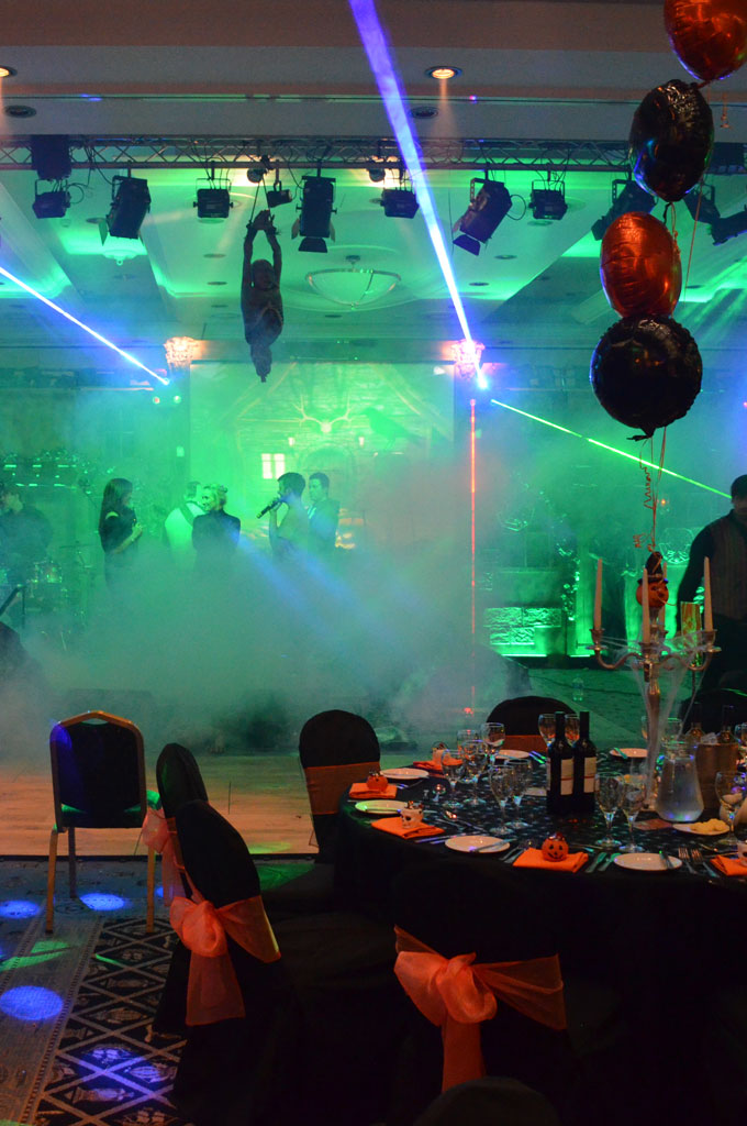 An example of corporate event production showing staff on stage singing karaoke with microphones in laser lighting and smoke effects, with balloons and a halloween decorated round table in the foreground.