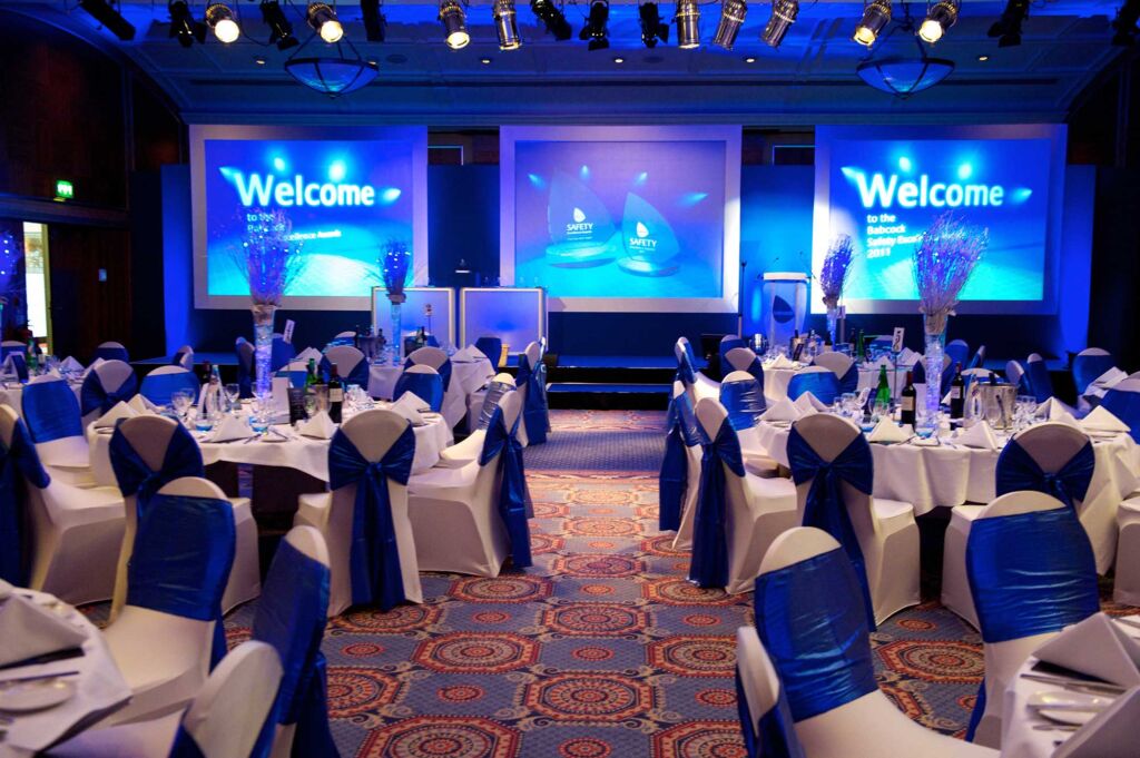 Babcock Annual Conference and Awards Ceremony with screens on the back wall and round tables with tall flower arrangements, chairs with fabric sashes, showing an example of corporate event production.