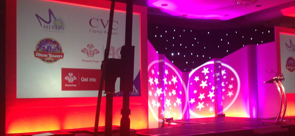 Example of conference stage design showing The Prince's Trust at Alton Towers event with LCD screens, stage with gobo lighting effects with stars and lectern, and close up of microphone stand.