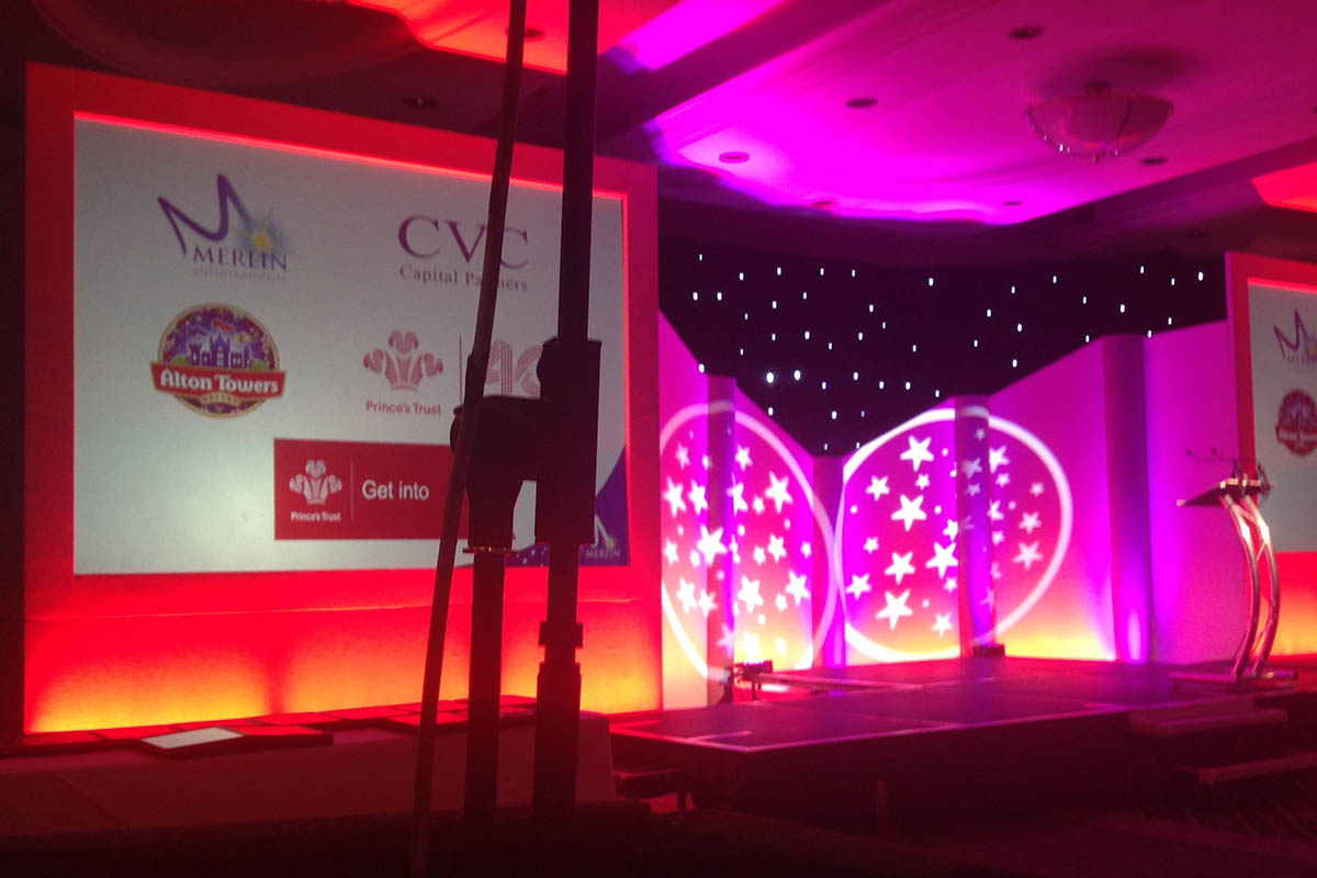 Example of conference stage design showing The Prince's Trust at Alton Towers event with LCD screens, stage with gobo lighting effects with stars and lectern, and close up of microphone stand.