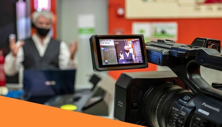 A man is being filmed by a camera, surrounded by other corporate event production equipment in a corporate office environment.