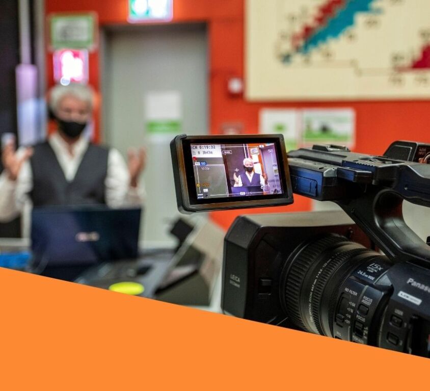 A man is being filmed by a camera, surrounded by other corporate event production equipment in a corporate office environment.