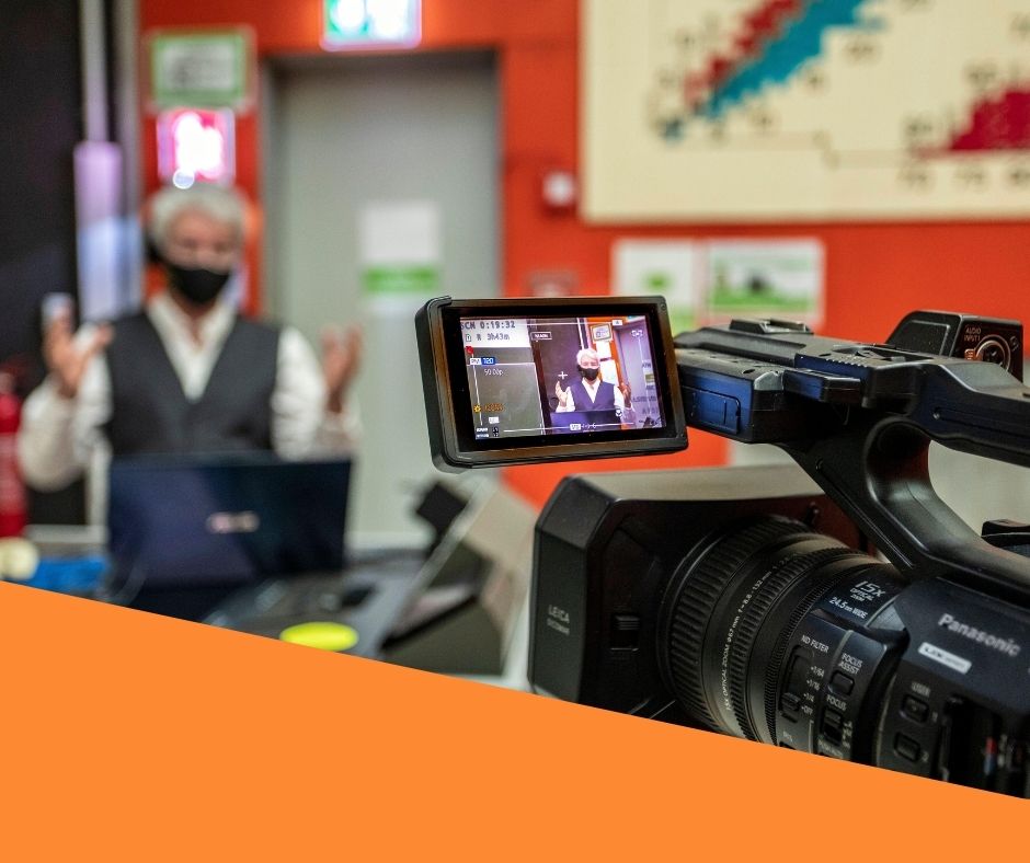 A man is being filmed by a camera, surrounded by other corporate event production equipment in a corporate office environment.