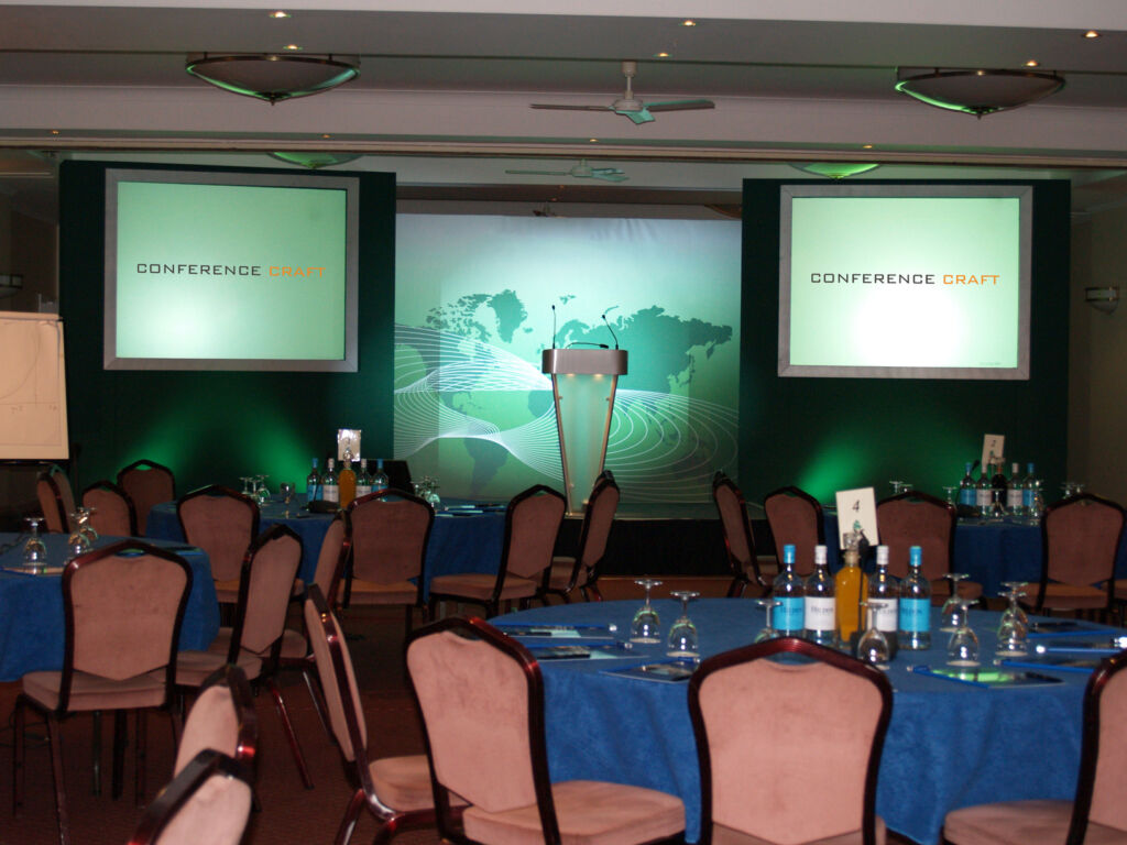 Round tables set up with chairs and drinks as for a conference, with a stage and lectern with screens showing "Conference Craft", who are a conference production company