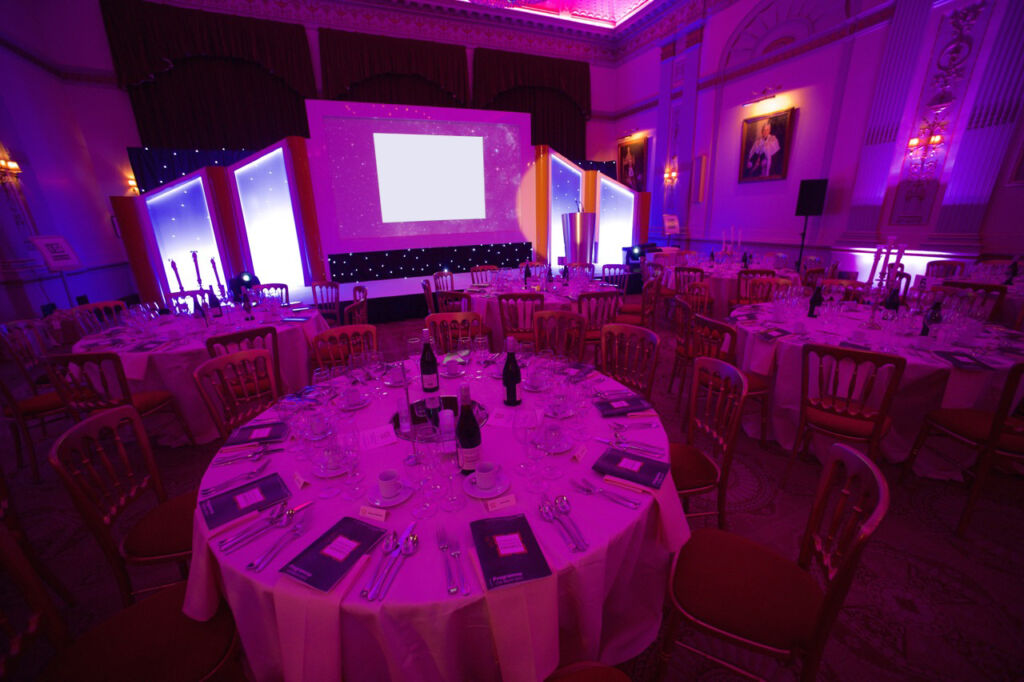 Circular tables set up for the UCLH Awards with drinks, cutlery, A4 packs of printed materials, lit as if evening with a pink lighting effect and a stage with LCD screens to illustrate corporate event production.