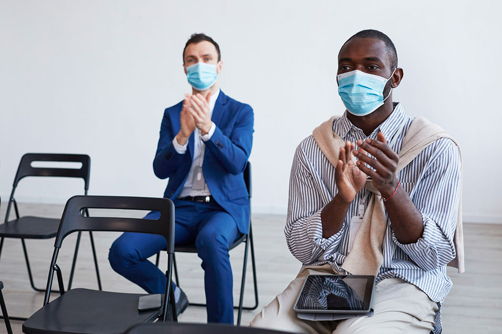 Two men sitting in conference seating applauding, socially distanced and wearing Covid masks, as might be organised by conference specialists in a post-pandemic world.