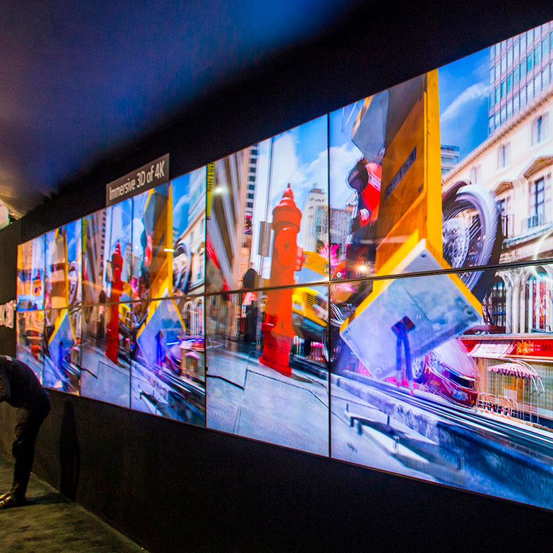 Conference stage design with a backdrop of an LCD wall displaying brightly coloured repeating images and a speaker in front of it.